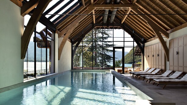Une piscine intérieure avec des poutres en bois, des parois en verre transparent et des chaises longues.