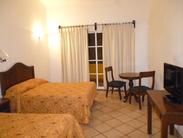 Habitación de hotel con dos camas, una mesita con sillas, televisor y ventana con cortinas.