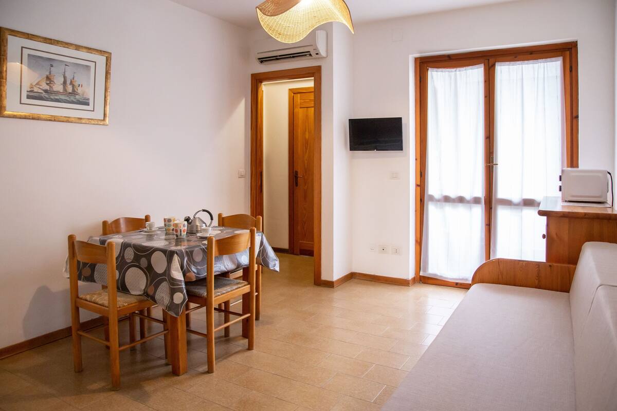 A dining area with a table and chairs, a television mounted on the wall, and a sliding glass door leading to a balcony.