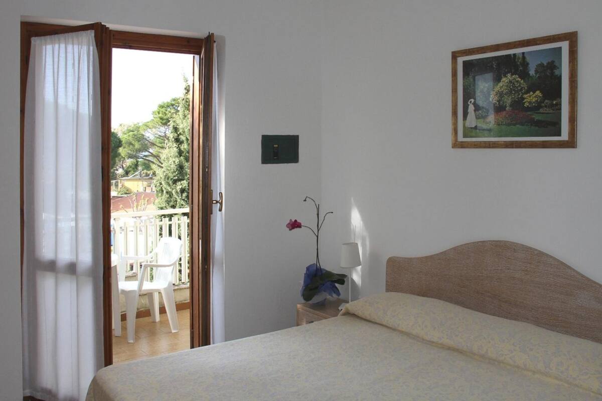 A bedroom with a bed, a nightstand, a window with curtains, a painting on the wall, and a view of the outside balcony.