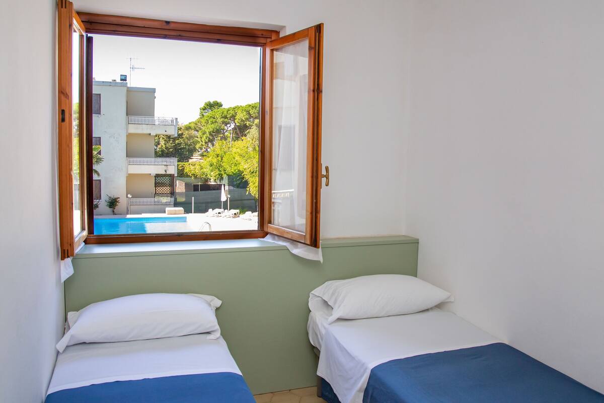 A room with two beds, a window with a view of a swimming pool and buildings, and a green wall.