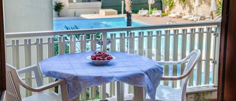 A balcony with a table set for two, a bowl of fruit, and a pool in the background.