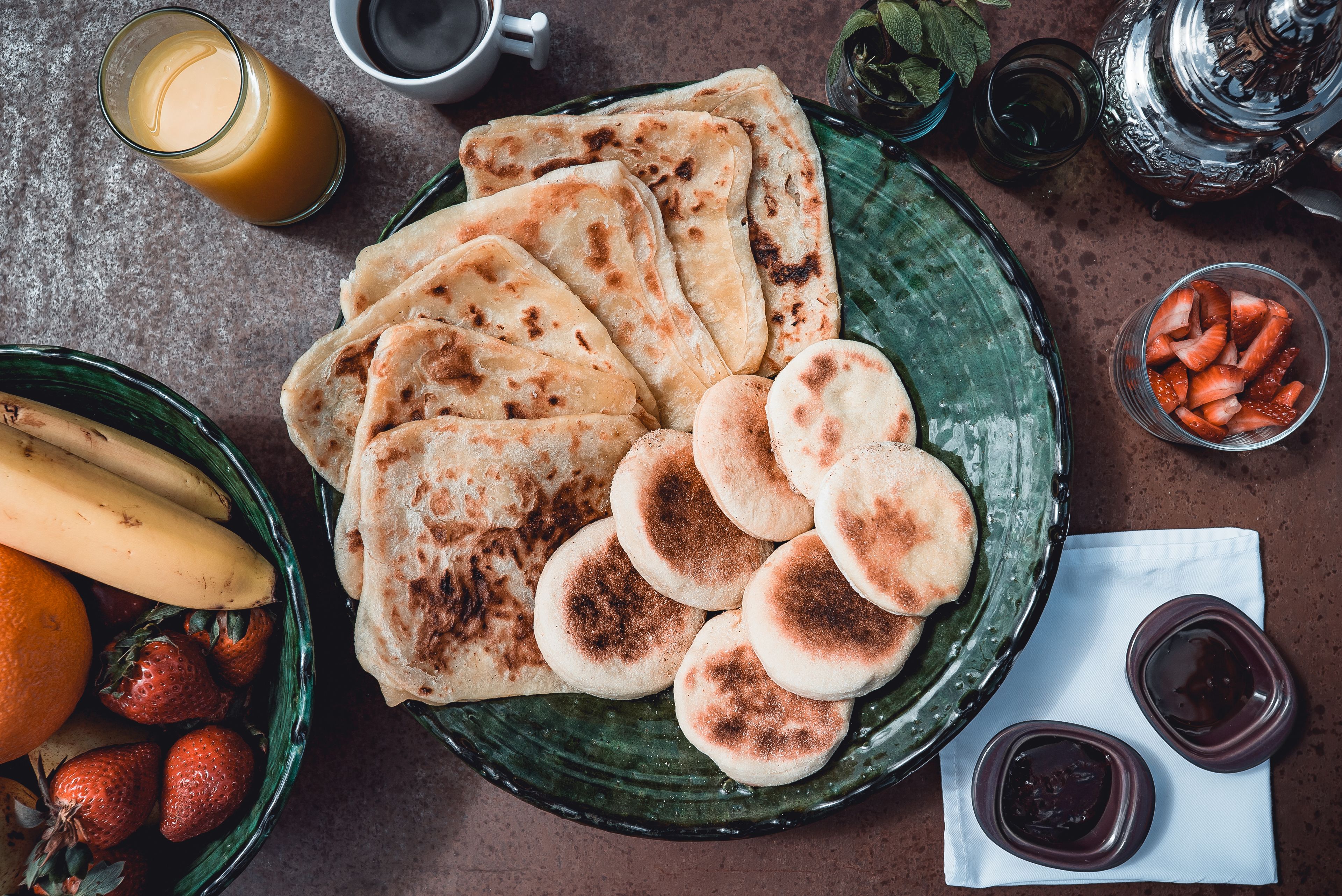 Un plat de flatbreads, un bol de maduixes, plàtans i un got de suc d'aranja sobre una taula.