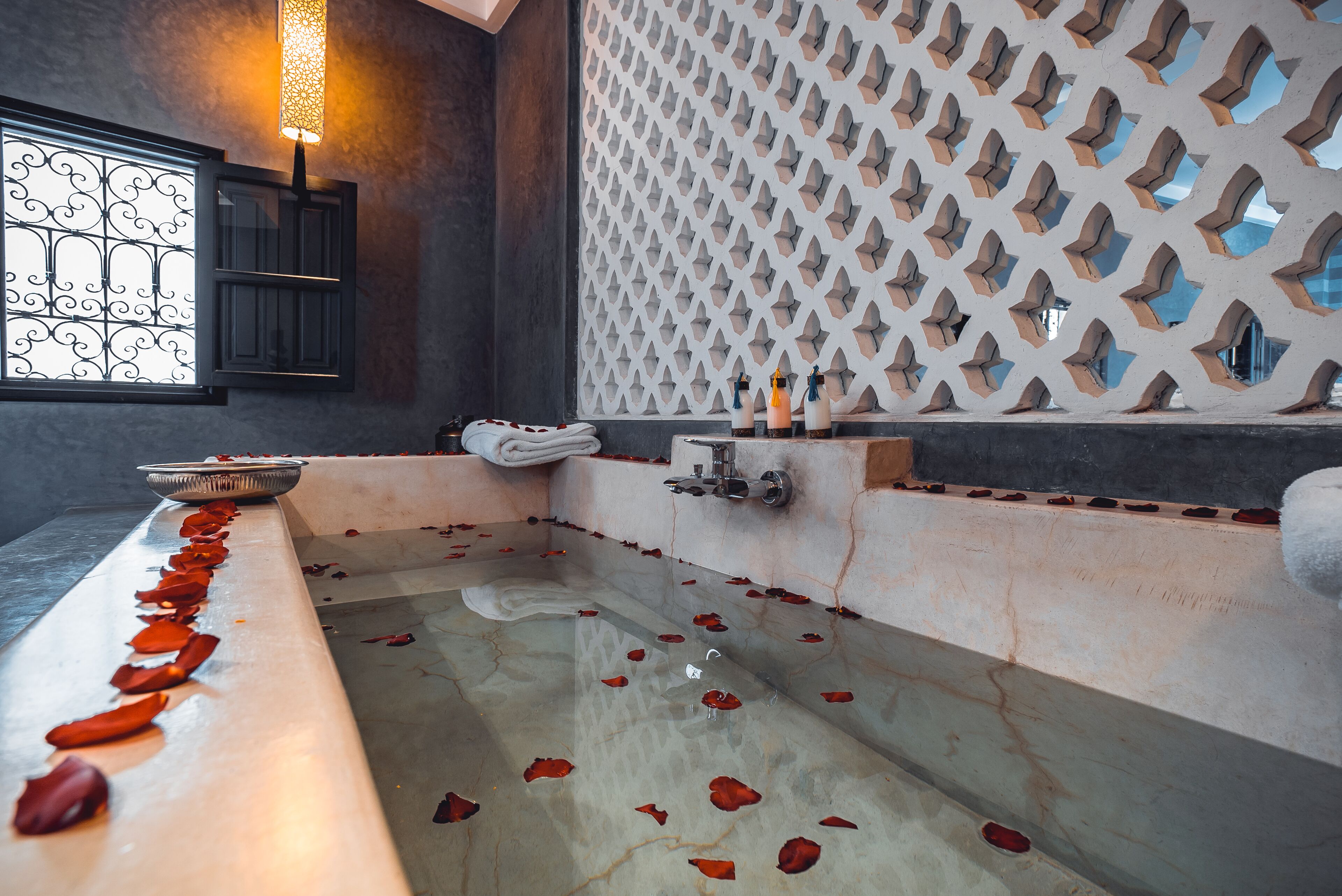 Una bañera con pétalos de rosa, un muro decorativo y una ventana con reja de metal.