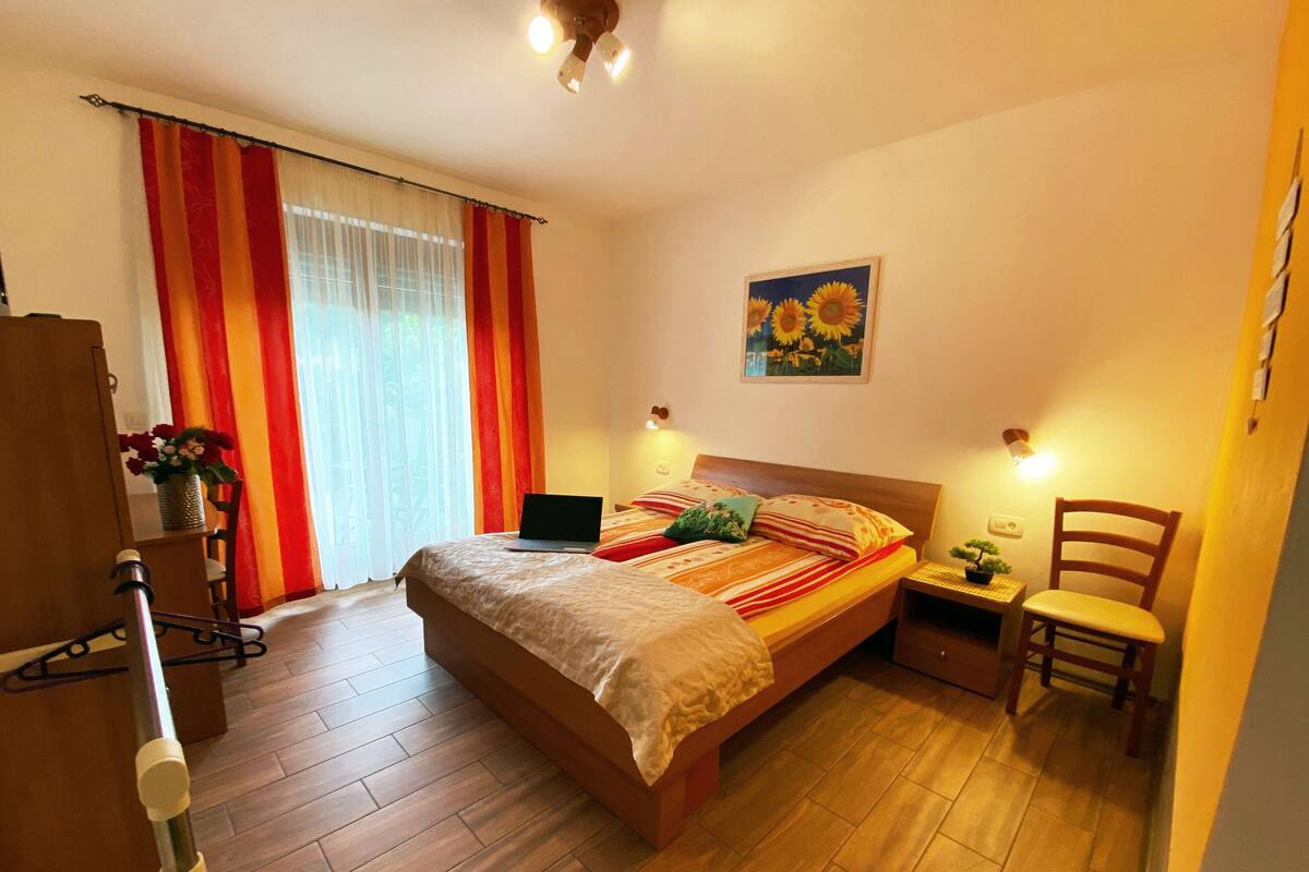 Een slaapkamer met een houten bed, een laptop op het bed, een stoel, een klein tafeltje met een plant en een schilderij aan de muur.