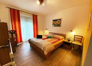Une chambre avec un lit en bois, un ordinateur portable posé sur le lit, une chaise, une petite table avec une plante et un tableau accroché au mur.