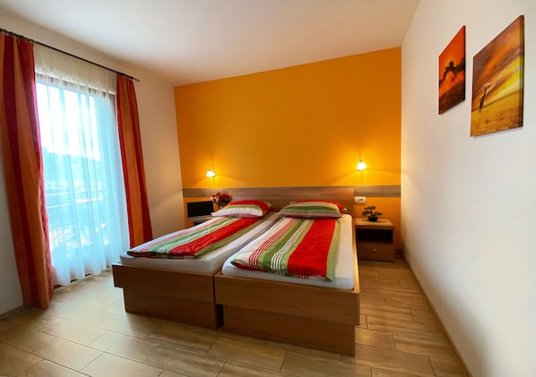 Une chambre d’hôtel avec deux lits, une grande fenêtre avec des rideaux, deux lampes fixées au mur et des œuvres d’art accrochées au mur.