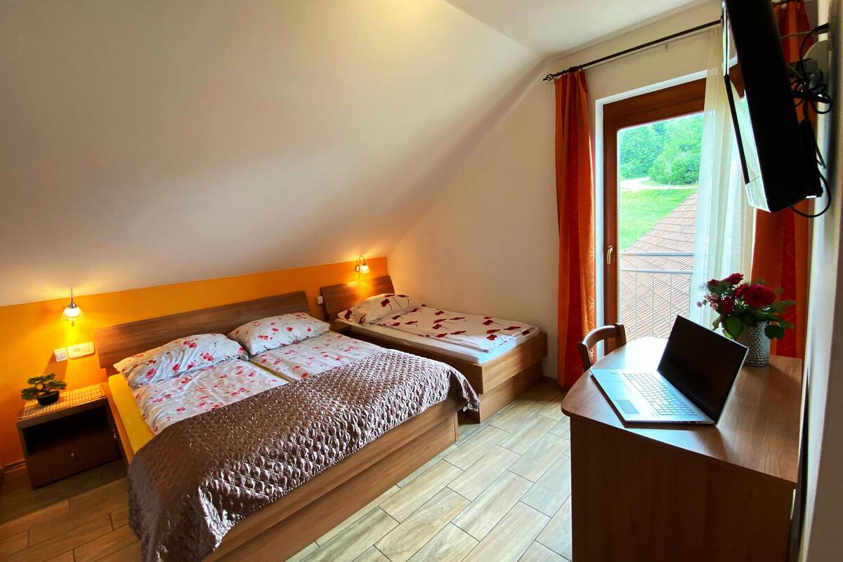 Een slaapkamer met een bed, een bureau met een laptop, een nachtkastje met een lamp en een raam met gordijnen.