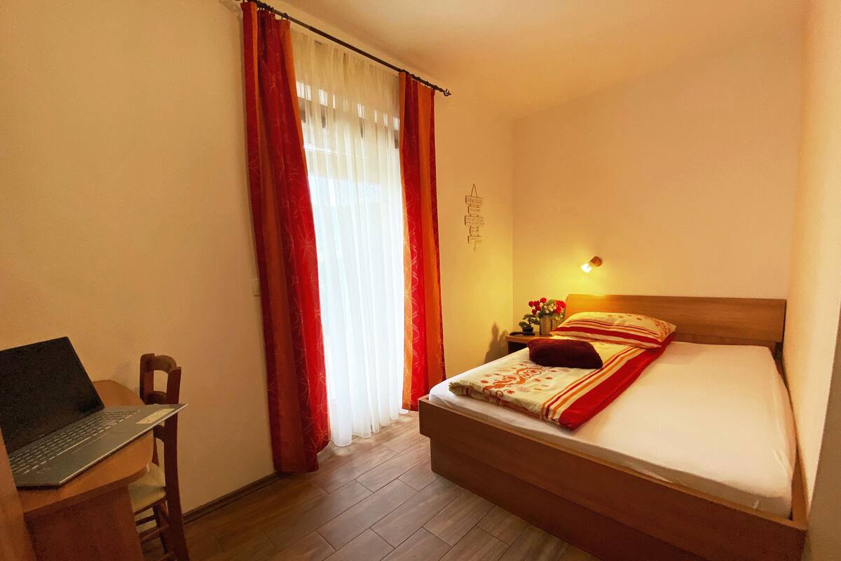 Hotelkamer met een bed, een bureau met een laptop, een nachtkastje met een lamp en een raam met gordijnen.