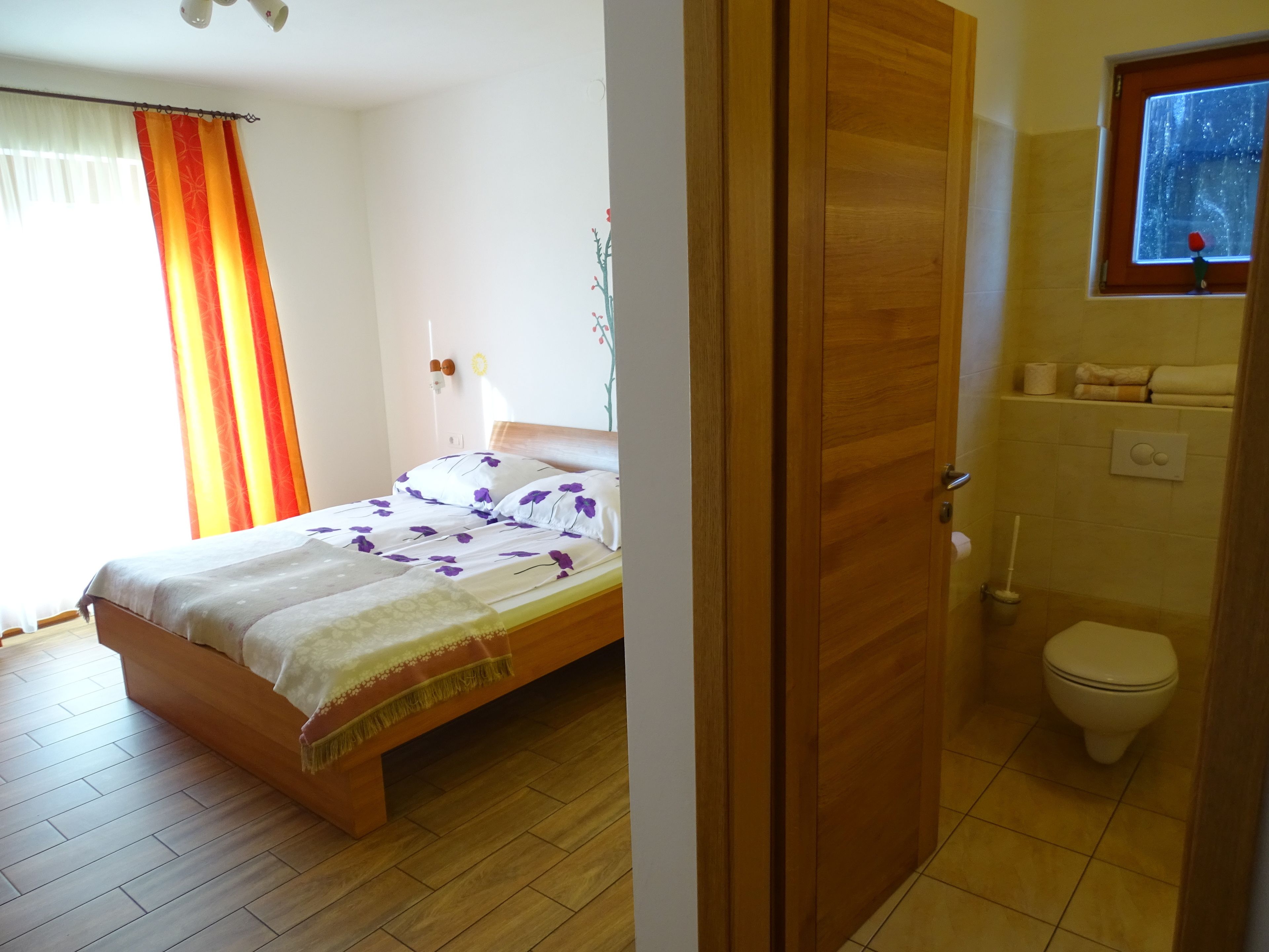 A bedroom with a wooden bed, a bathroom with a toilet, and a window with a view of the sky.