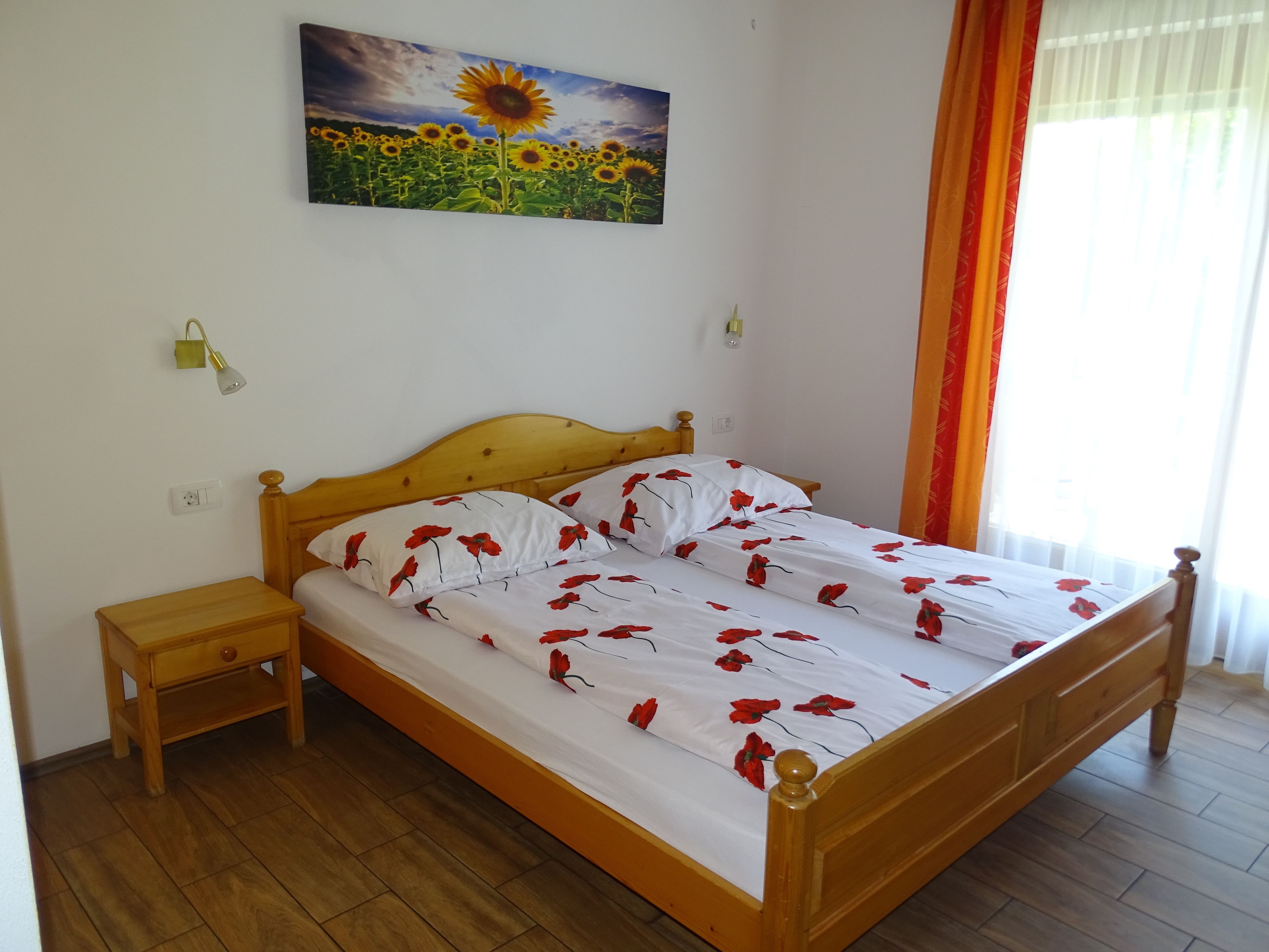 Ein Schlafzimmer mit einem hölzernen Bett, weißer Bettwäsche mit roten Blumenmustern, einem Nachttisch, einer Wandlampe und einem Gemälde von Sonnenblumen.