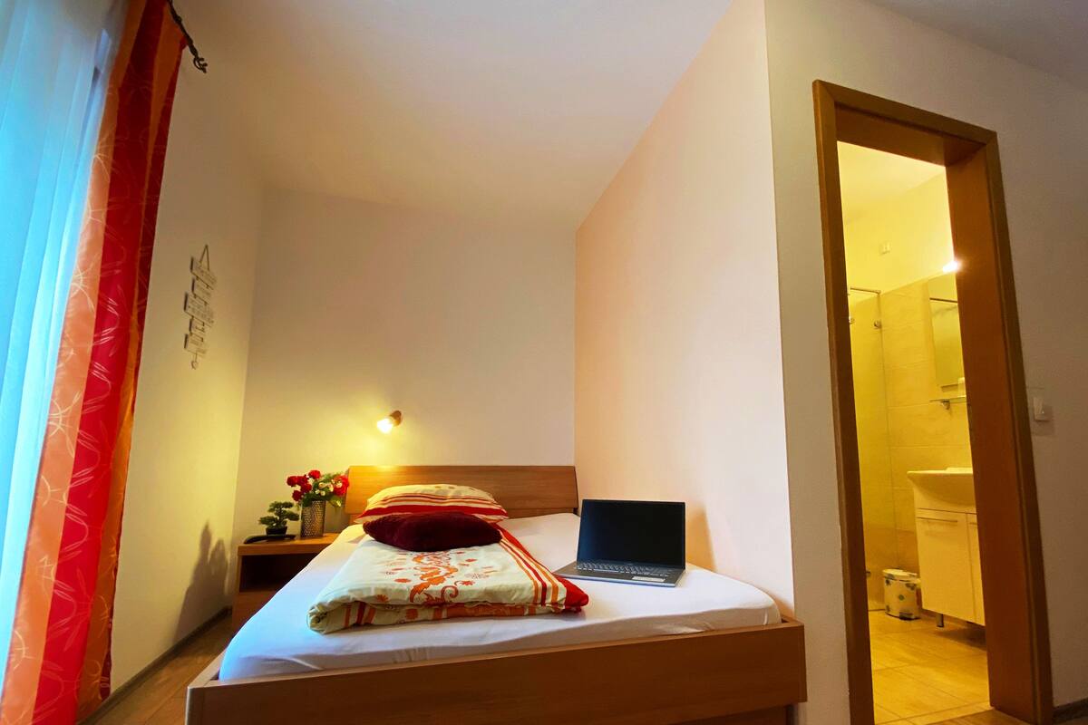 Een keurig opgemaakt bed met een laptop, een nachtkastje met een bloemetjesvaas, en een rood kussen.