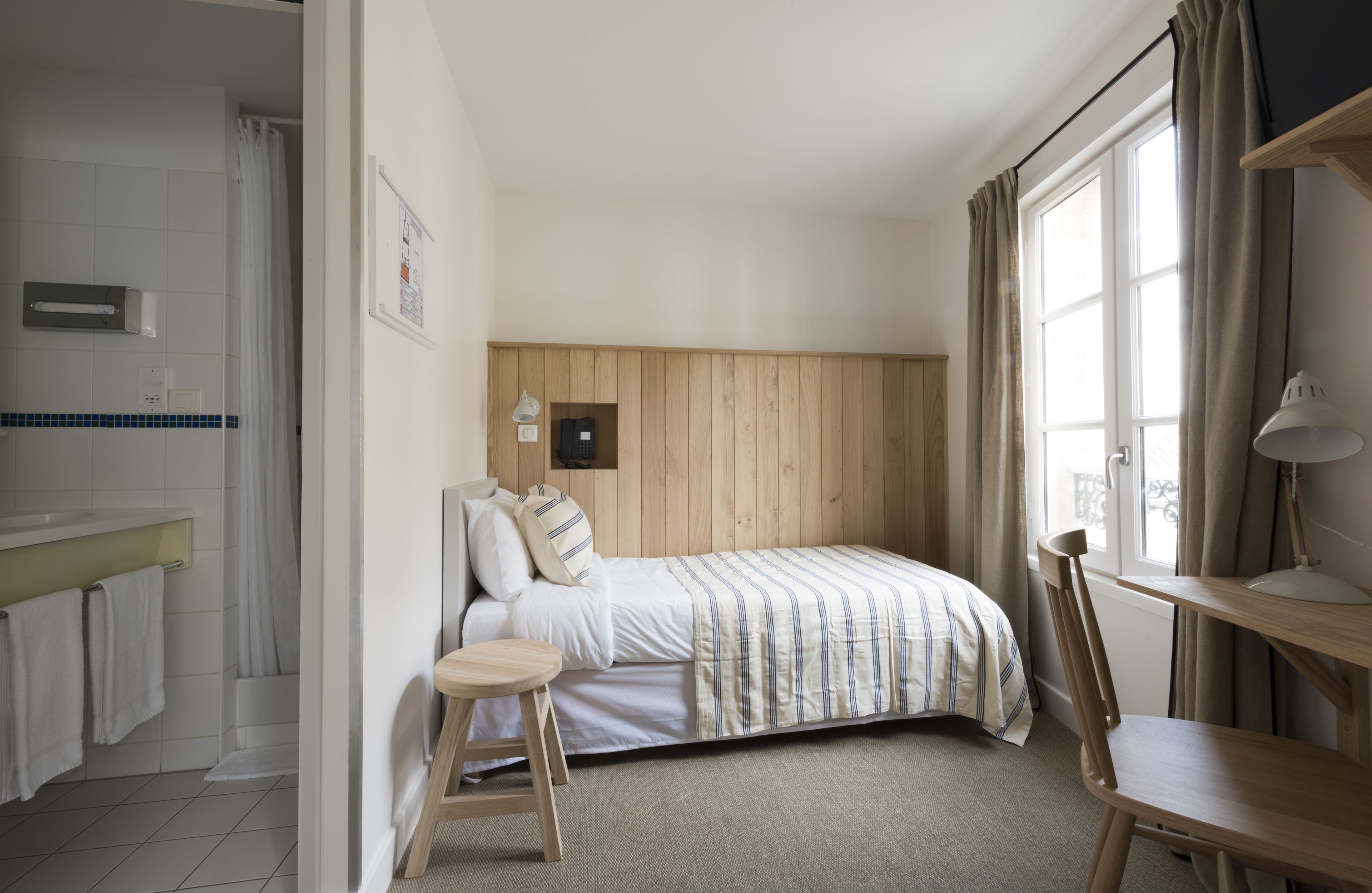 A bedroom with a bed, a wooden chair, a small stool, a desk with a lamp, and a window with curtains.