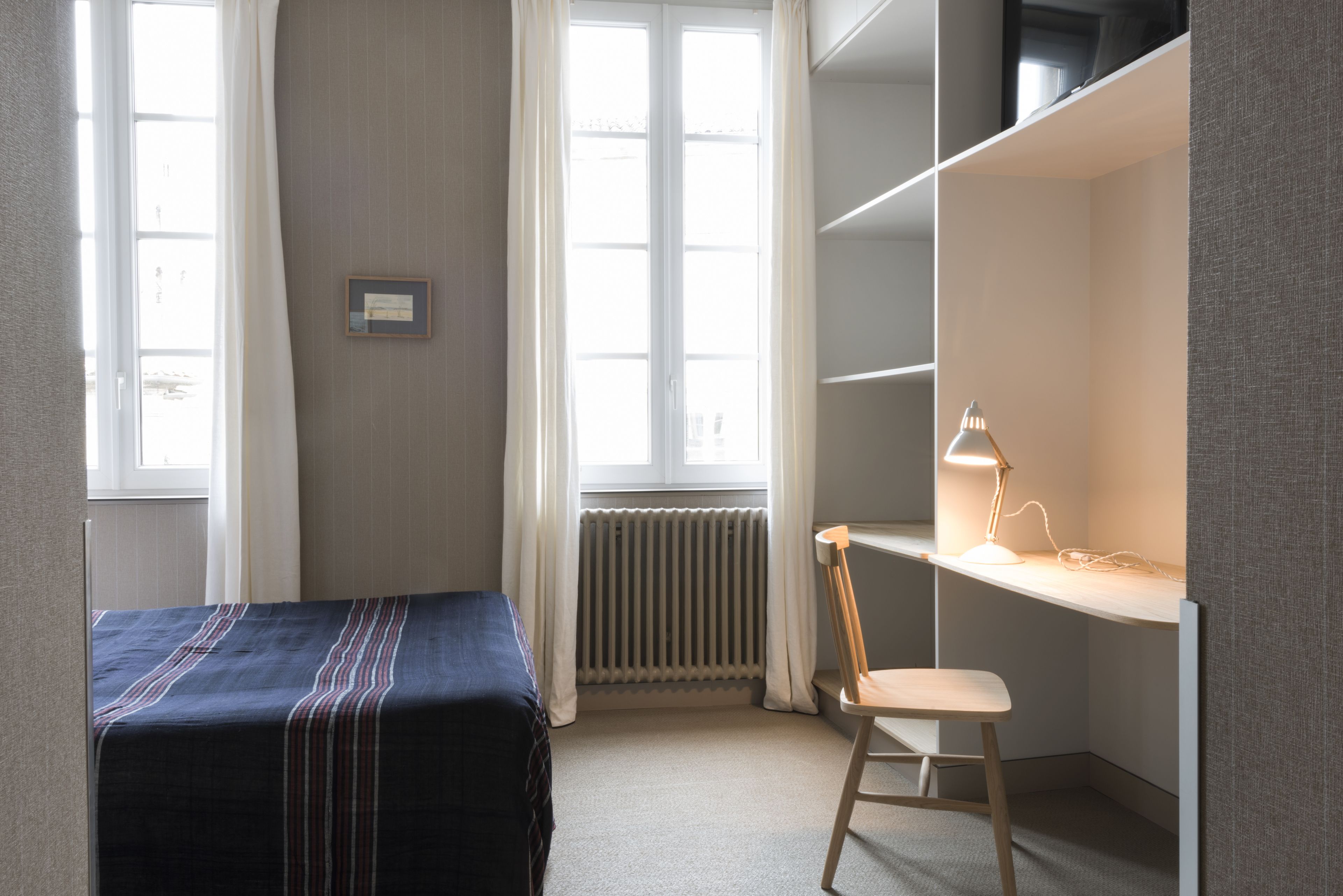 A bedroom with a bed, a desk with a chair, a radiator, and a window with curtains.