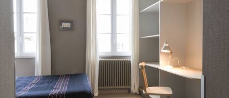 A bedroom with a bed, a desk with a chair, a radiator, and a window with curtains.