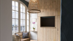 A room with a wicker sofa, a wooden coffee table, a TV mounted on a wooden wall, and a large window with multiple panes. - Auberge de la Commanderie (Saint-Émilion)