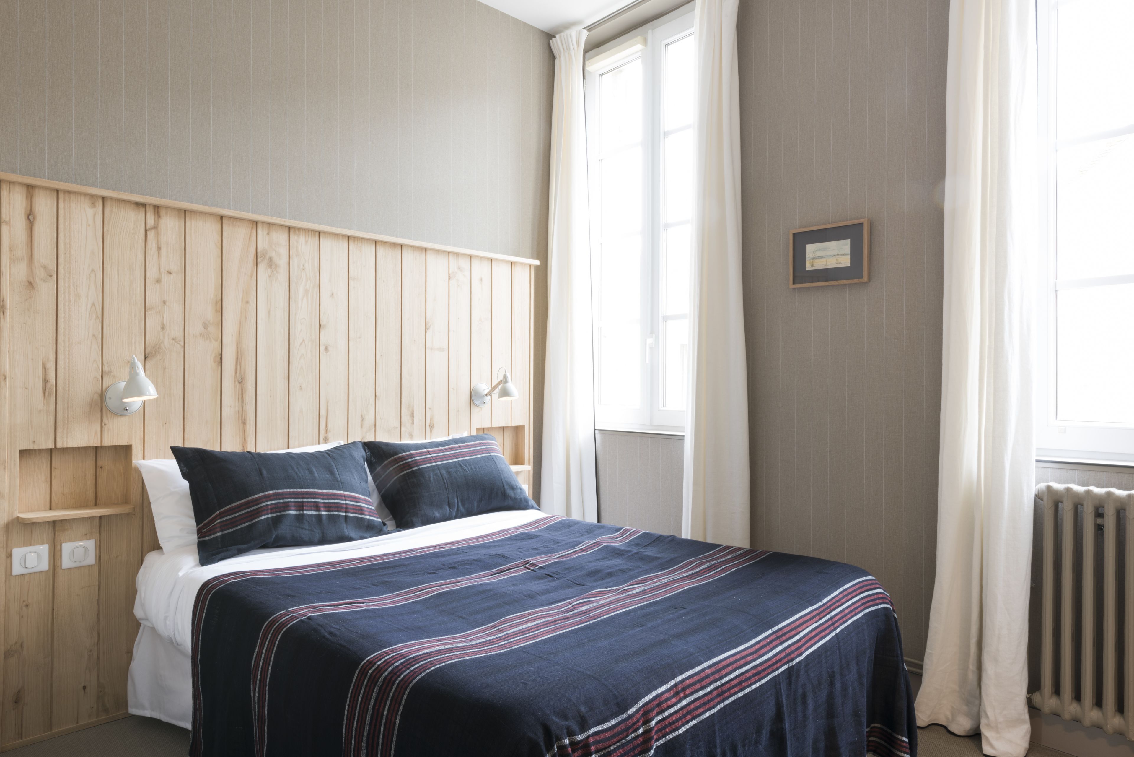 A bedroom with a wooden headboard, a bed with a striped bedspread, a bedside lamp, and a window with curtains.