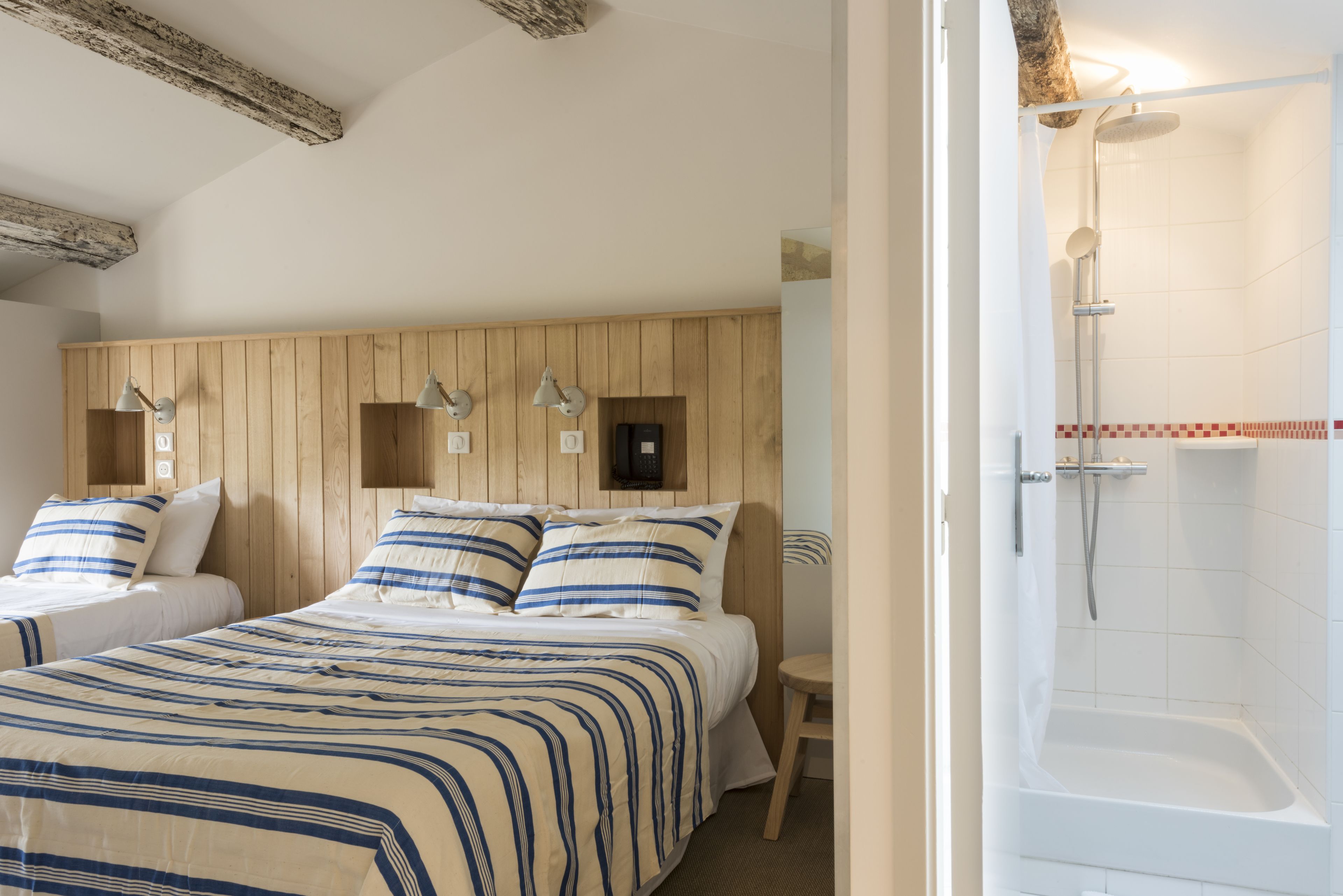 Habitación de hotel con cabecera de madera, una cama grande con ropa de cama a rayas, una mesita de noche pequeña y una zona de ducha con bañera blanca.