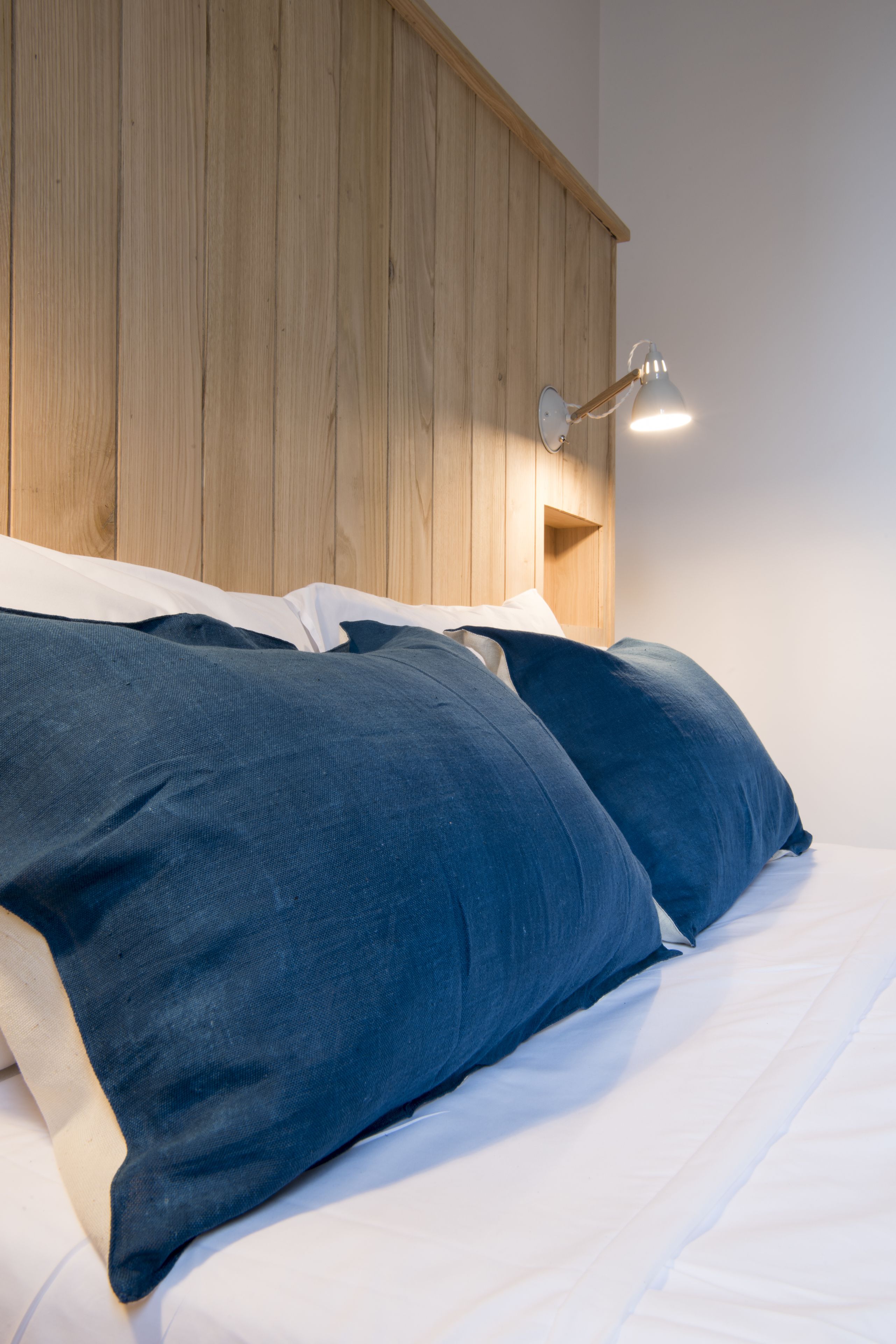 Un letto con lenzuola bianche e due cuscini blu scuro, una testiera in legno e una lampada fissata al muro.