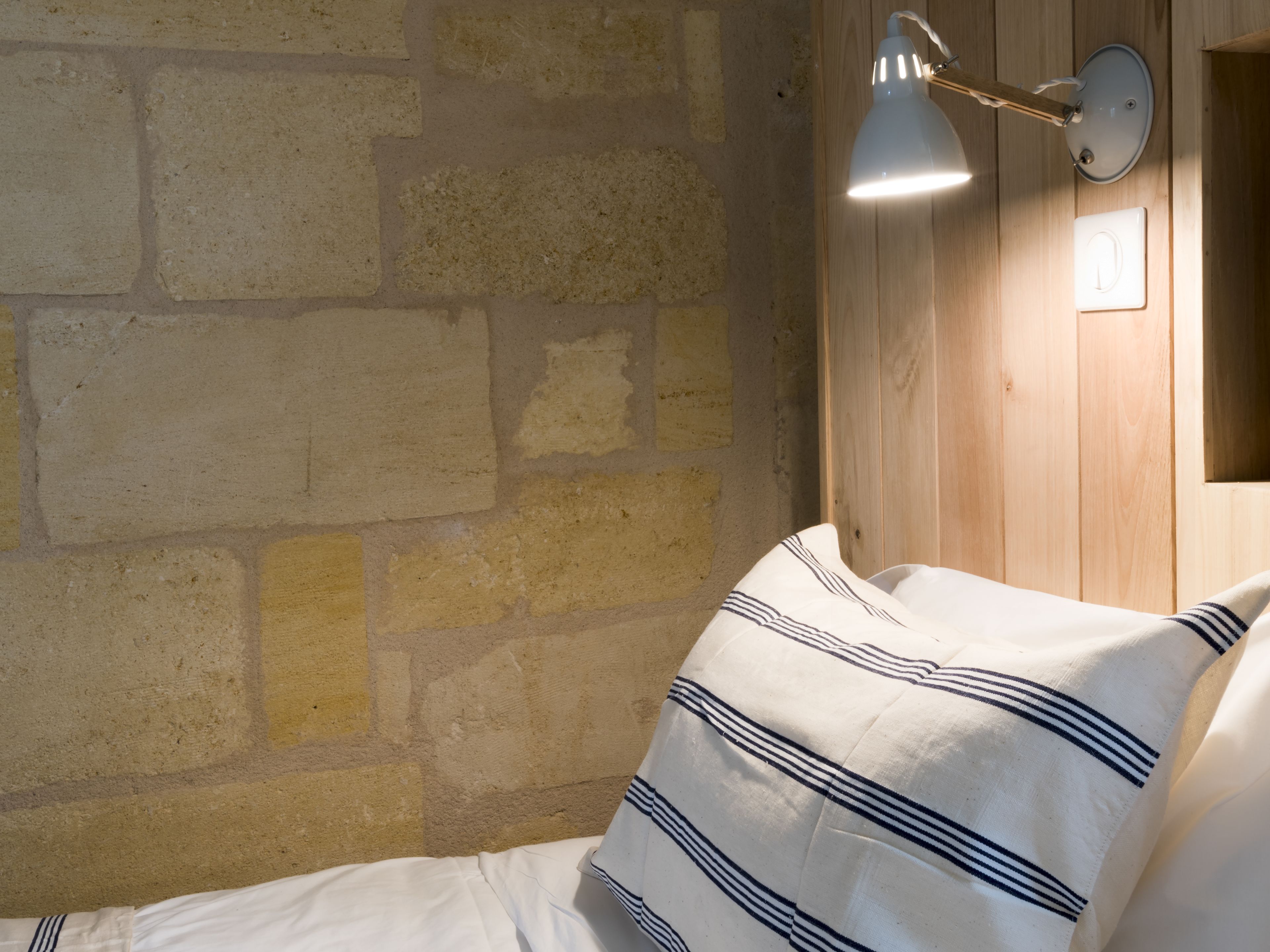 Una habitación con pared de ladrillo, pared de paneles de madera, cama con ropa blanca y almohadas a rayas, y una lámpara fijada a la pared.