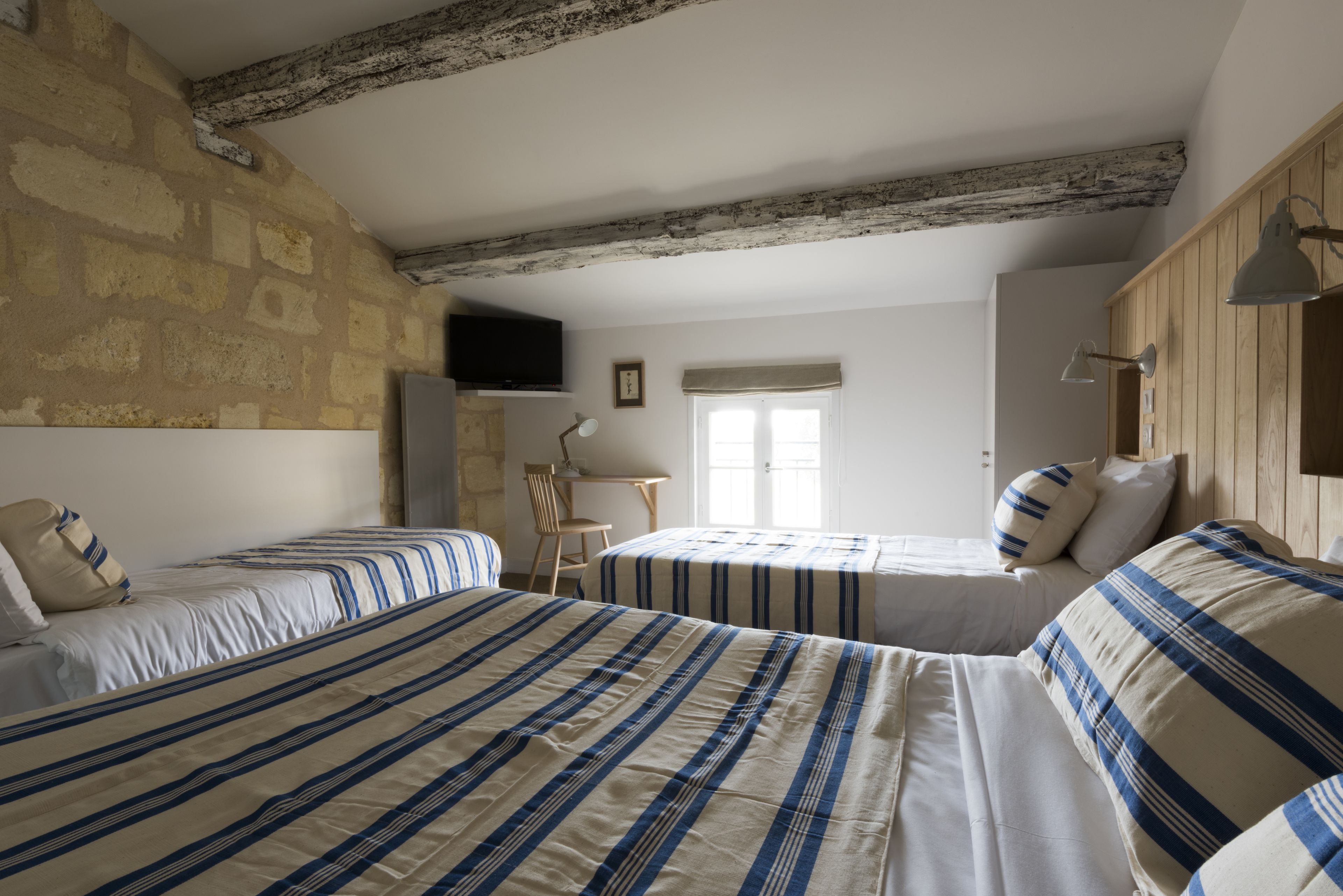 A room with two beds, a wooden ceiling, a television on the wall, and a window with curtains.