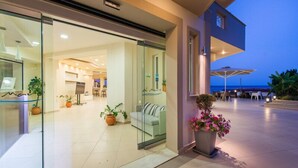 Un moderno vestíbulo de hotel con entrada de vidrio, una zona de estar con sofá y sillas, y un balcón con mobiliario exterior.