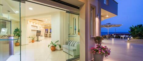 A modern hotel lobby with a glass entrance, a seating area with a sofa and chairs, and a balcony with outdoor furniture.