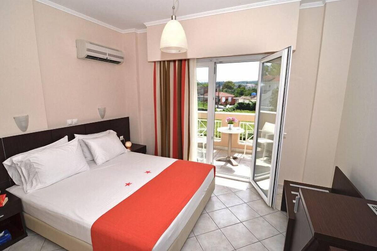 A hotel room with a bed, a nightstand, a desk, a chair, a balcony with a table and chairs, and a view of the outside.