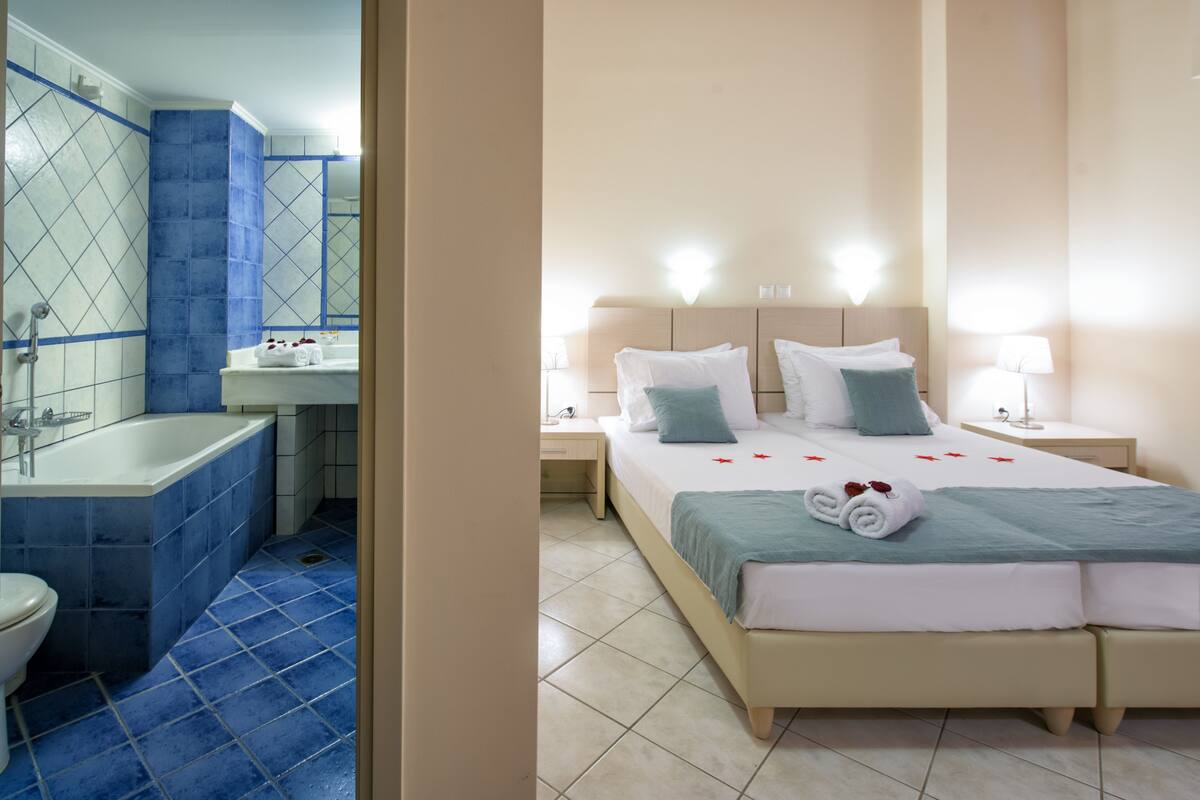 A hotel room with a bathroom, a bed with pillows, a nightstand, and a lamp.