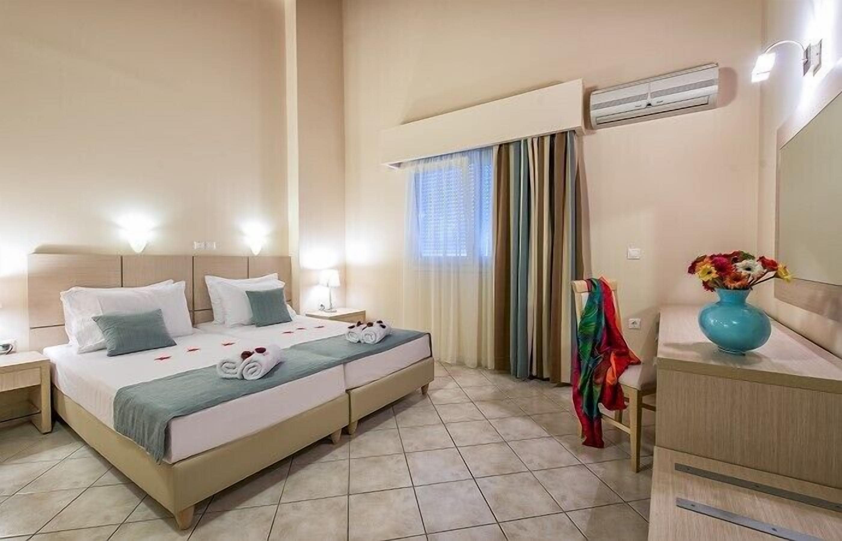 Ein Hotelzimmer mit Doppelbett, Nachttisch mit Lampe, kleinem Tisch mit Blumenvase und einem Fenster mit Vorhängen.