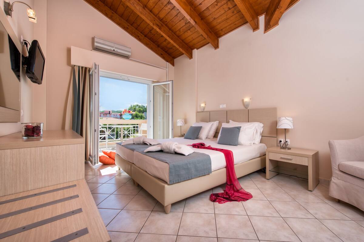 A bedroom with a bed, bedside tables, a nightstand, a TV, and a window with a view of a pool and buildings.
