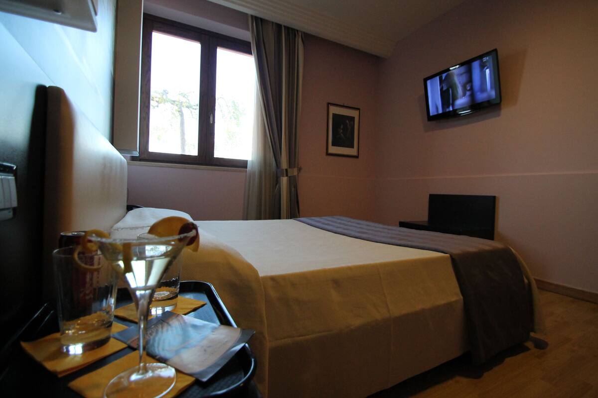 A hotel room with a bed, a TV mounted on the wall, a window with curtains, and a table with glasses and a tray.