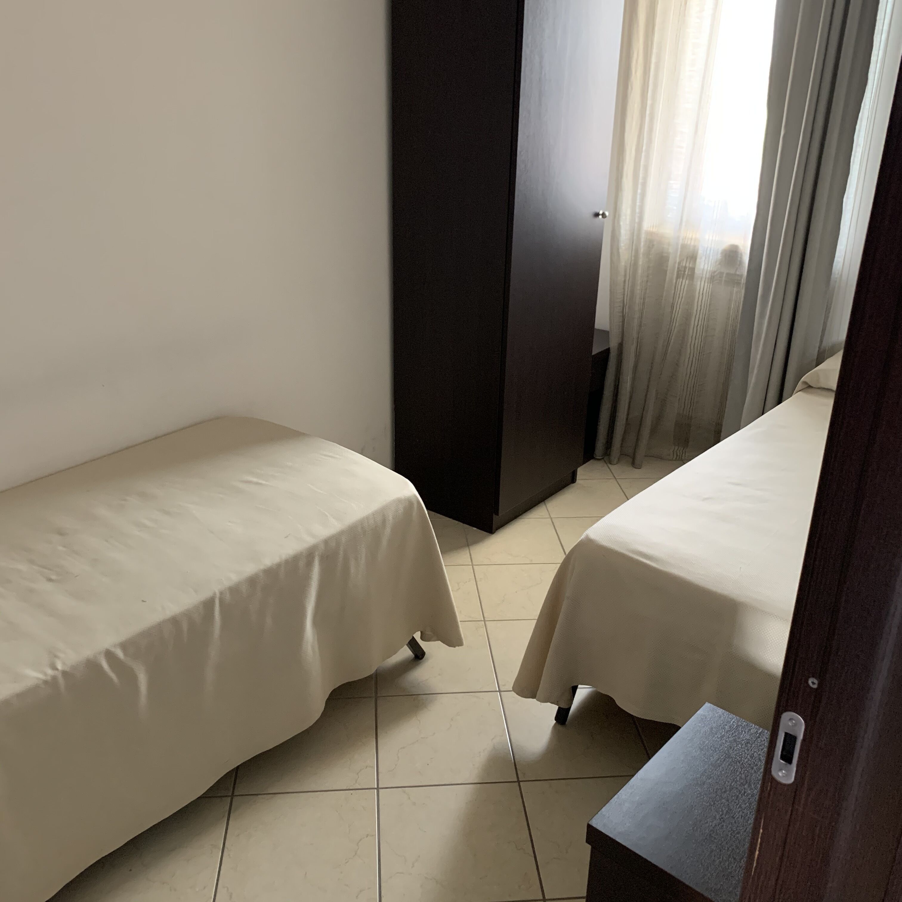 A small, clean room with two single beds, a dark-colored wardrobe, and a window with curtains.