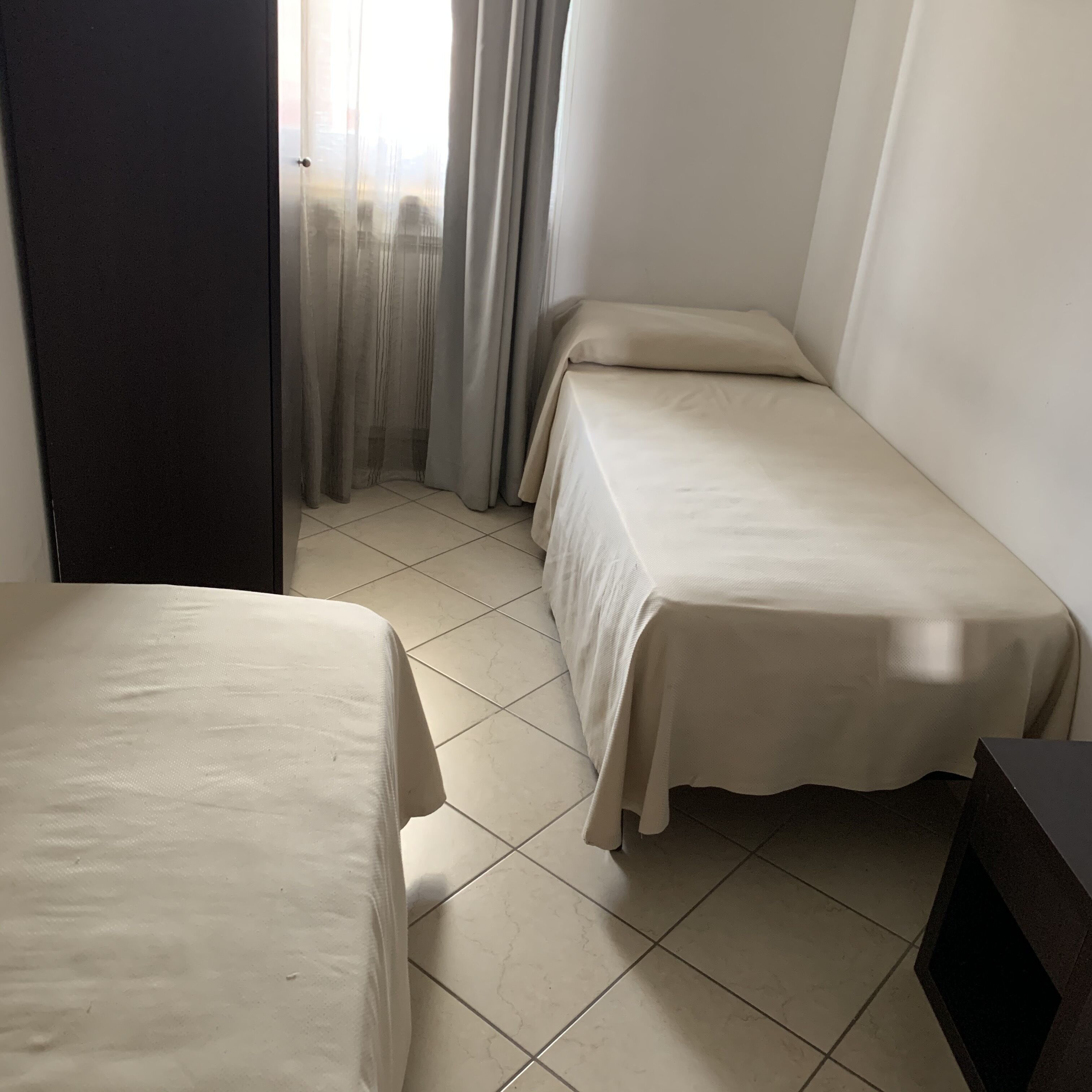 A small, clean room with two single beds, a tiled floor, and a door leading to another room.