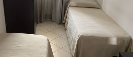 A small, clean room with two single beds, a tiled floor, and a door leading to another room.