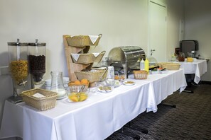Disposição de buffet com diversos alimentos e bebidas, incluindo um dispensador de café, cestas e um jarro de suco.