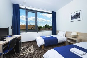 Quarto de hotel com duas camas, uma escrivaninha, uma cadeira, janela com cortinas, radiador e um quadro na parede.