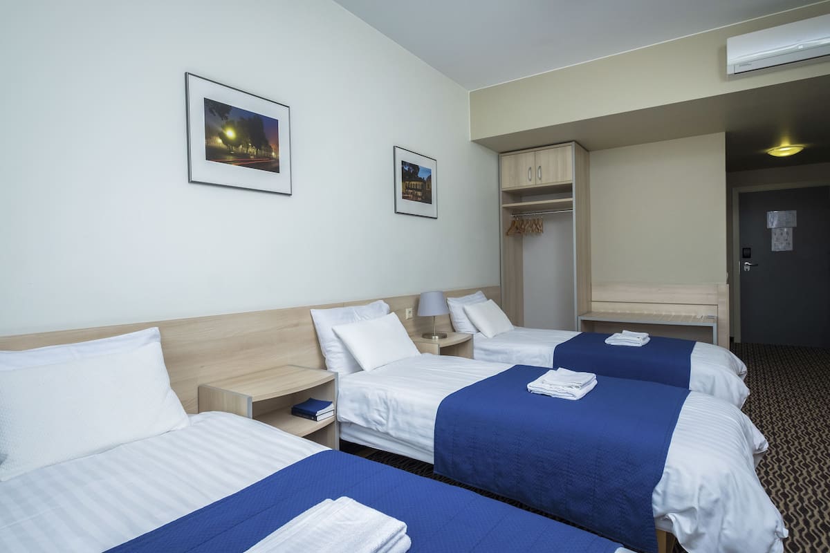 Habitación de hotel con tres camas, un armario, una mesita de noche con lámpara y cuadros en la pared.