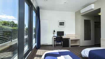 Une chambre d’hôtel avec deux lits, un bureau équipé d’un ordinateur, une chaise et une vue sur la verdure grâce à de grandes fenêtres.