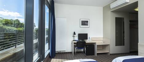 Une chambre d’hôtel avec deux lits, un bureau équipé d’un ordinateur, une chaise et une vue sur la verdure grâce à de grandes fenêtres.