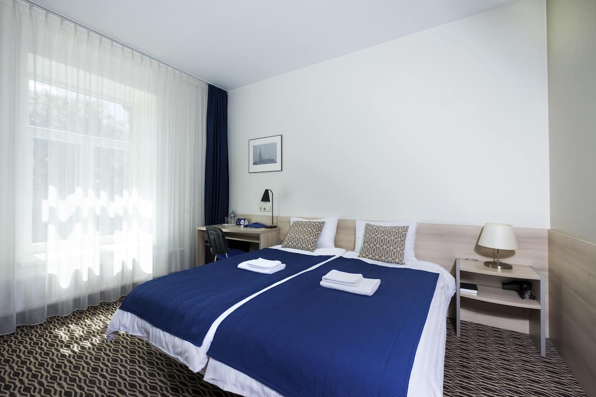 Habitación de hotel con una cama grande, un escritorio, una silla, una mesita de noche y una ventana con cortinas translúcidas.