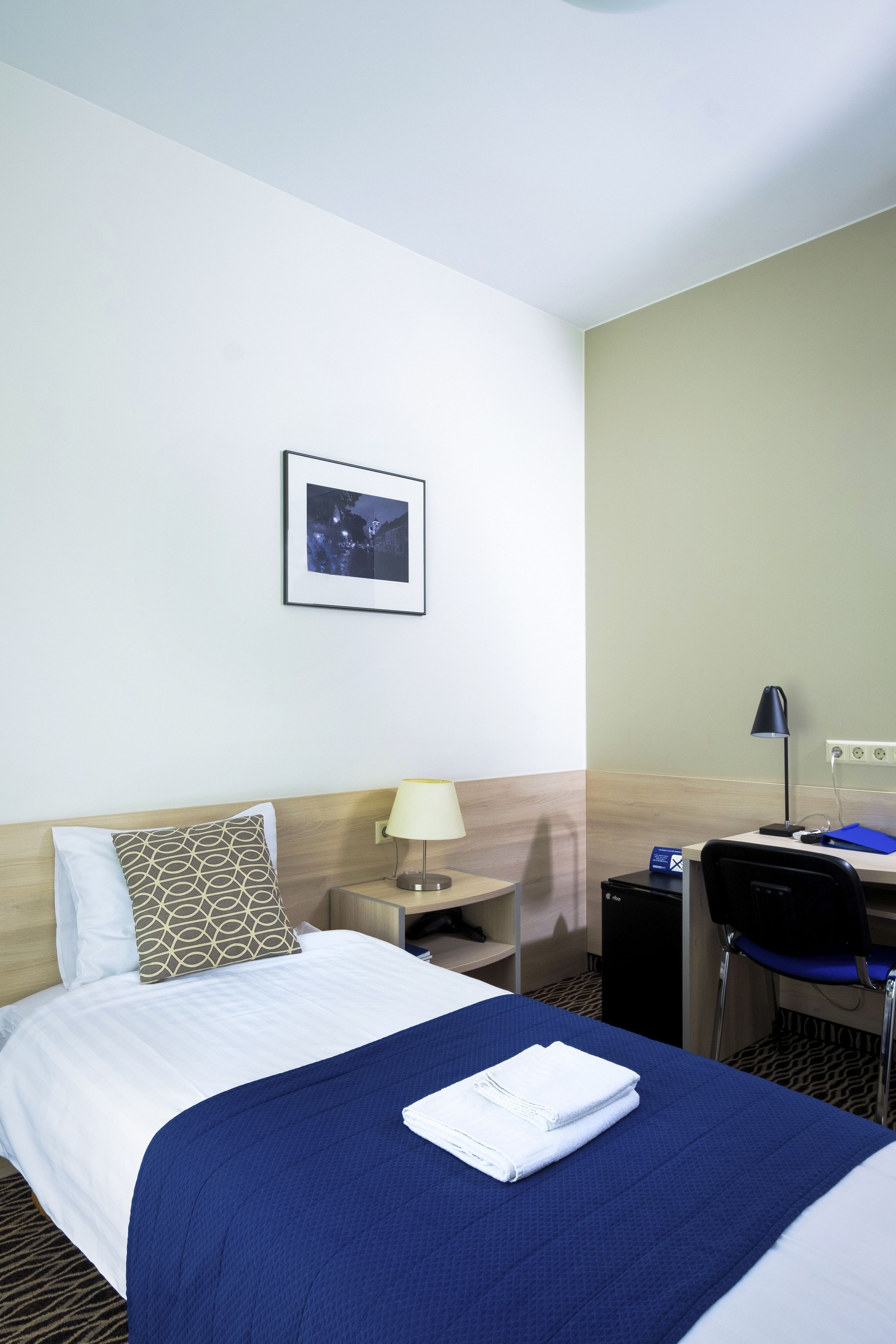 Una habitación de hotel con cama, escritorio, silla, lámpara y un cuadro en la pared.