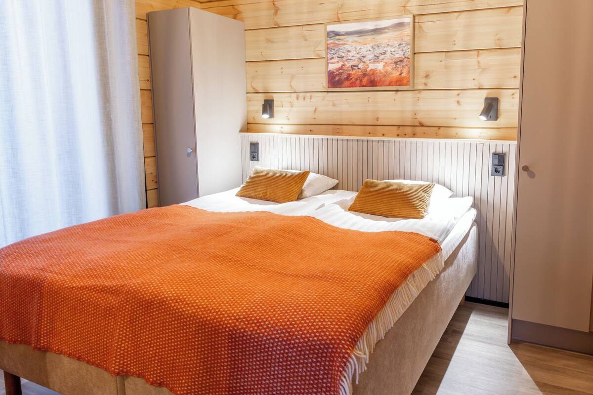 A room with a bed, wooden panel walls, a painting, and a nightstand with a lamp.