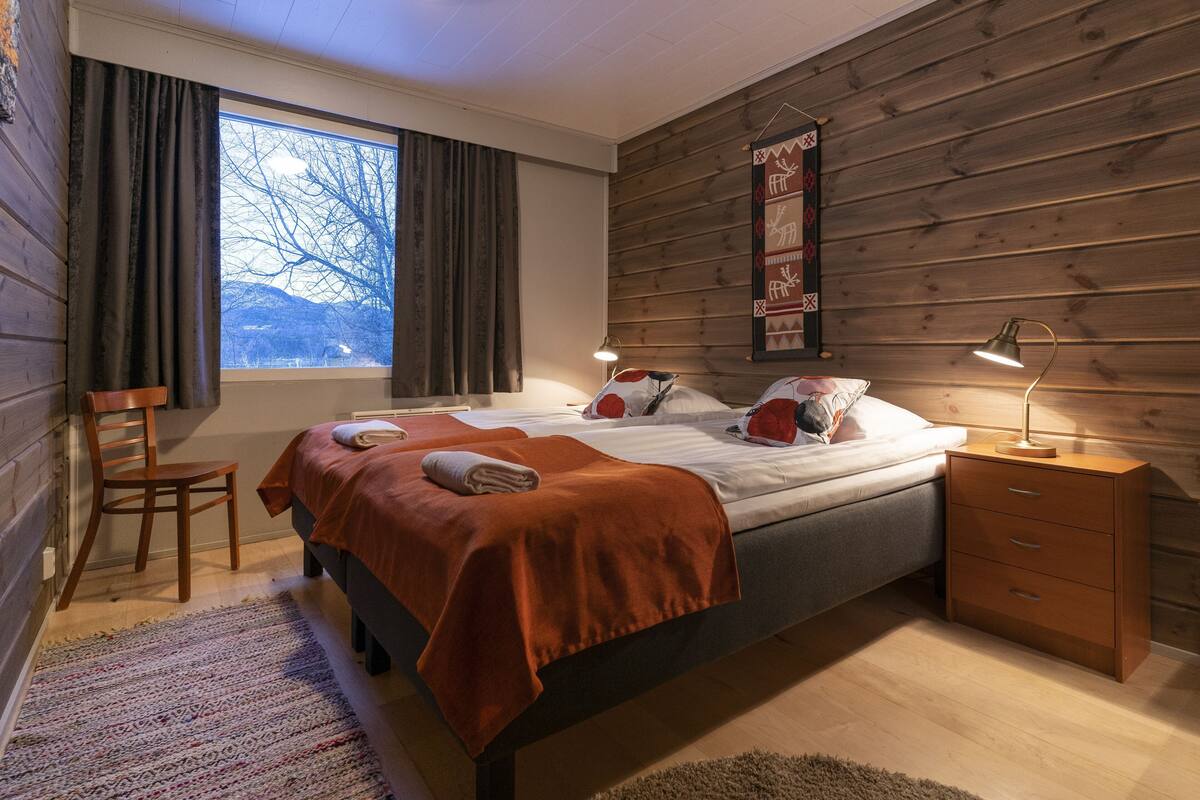 A bedroom with a bed, a nightstand, a chair, a window with curtains, and a wooden wall.