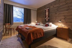 Un dormitorio con pared de madera, un ventanal grande, una cama con manta roja, una silla y una mesita de noche con una lámpara.