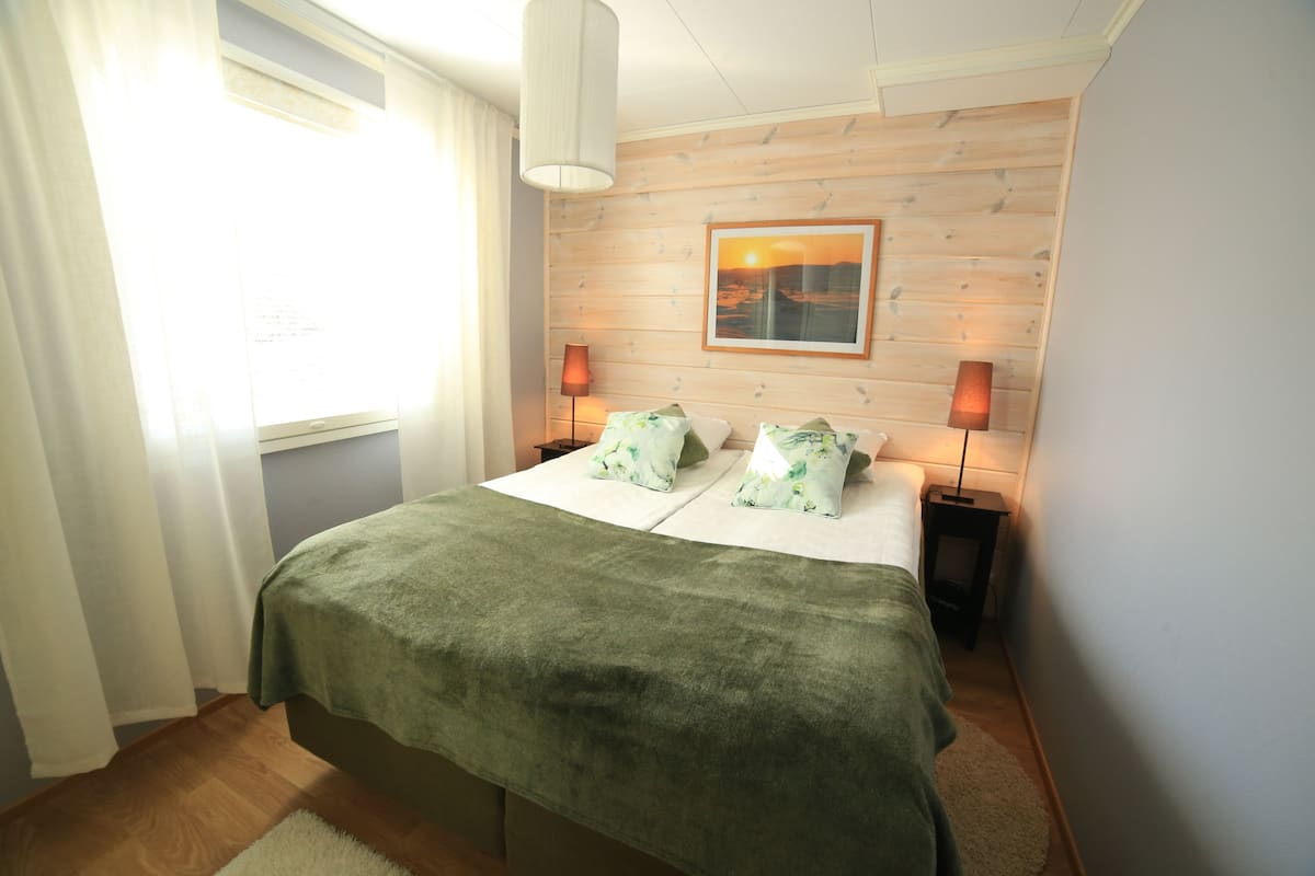 A bedroom with a bed, two bedside lamps, a window with curtains, a wooden wall, and a framed picture.