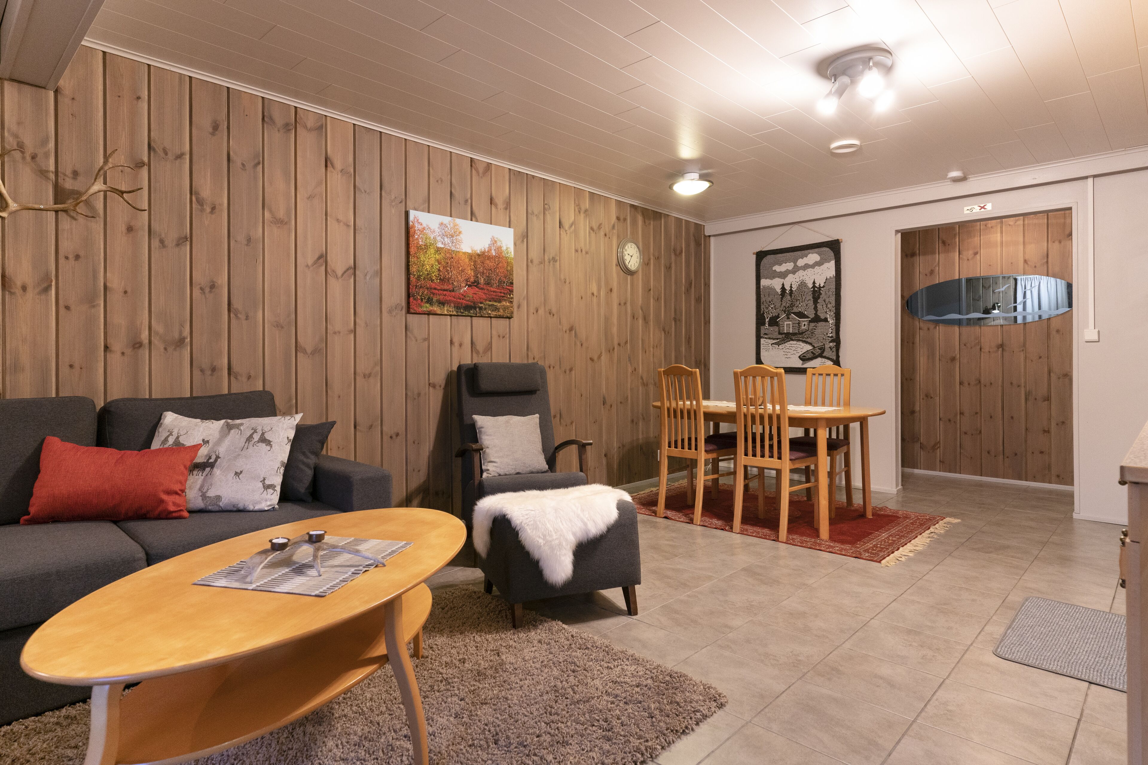 A modern living room with a wooden paneled wall, a grey sofa, a wooden coffee table, and a dining area with wooden chairs and a red rug.