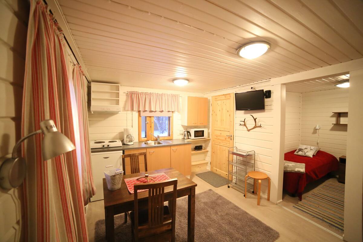 A compact kitchen with a dining area, a TV mounted on the wall, and a small seating area with a red bed.