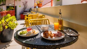 A table with breakfast items including croissants, ham, and a glass of orange juice, set on a balcony with potted plants and yellow chairs in the background. - Casa do Bairro by Shiadu (Lisbon)