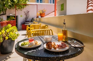 Uma mesa com itens de café da manhã, incluindo croissants, presunto e um copo de suco de laranja, em uma varanda com plantas em vasos e cadeiras amarelas ao fundo.